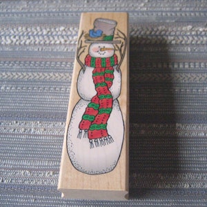 May include: Wooden stamp featuring a snowman wearing a red and green striped scarf and a brown hat with a blue bird on top.