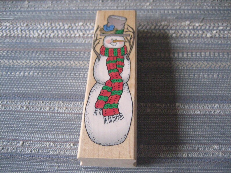 May include: Wooden stamp featuring a snowman wearing a red and green striped scarf and a brown hat with a blue bird on top.