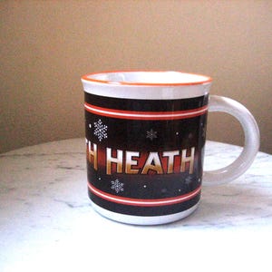 May include: A white ceramic mug with a black band featuring the name "Heath" in gold lettering, snowflakes, and red accents. The mug has a white handle and a white interior with an orange rim. The mug is on a white marble surface.
