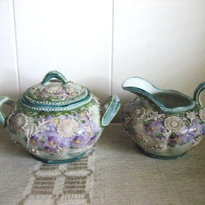 May include: A ceramic tea set featuring a teapot with a lid and a creamer. The set is decorated with a floral pattern in shades of purple, white, and green, with teal accents. The teapot has a handle and a knob on the lid.