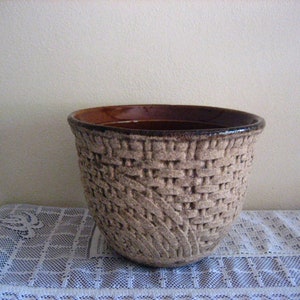 May include: A beige ceramic basket-style planter with a woven texture and a brown interior. The planter has a wide, open top and is suitable for indoor or outdoor use. The planter is sitting on a white lace tablecloth.