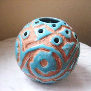 May include: A spherical ceramic vase with a turquoise glaze and brown carved details. The vase has several circular openings and a textured surface. The design features a repeating pattern of carved shapes. This decorative vase is suitable for home decor.
