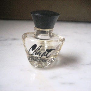 May include: A small, clear glass perfume bottle with a black cap. The bottle has a curved shape and features the text "Climat de Lancome" in black script. The bottle is sitting on a white surface.