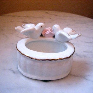 May include: A white ceramic trinket dish with a gold trim, featuring two white doves perched above a pink rose. The dish has a circular opening and is likely used for small jewelry or decorative items.