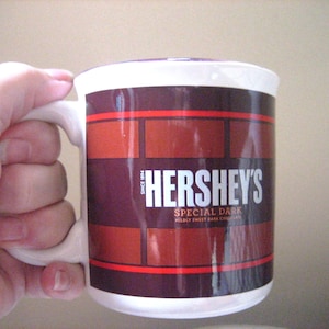 May include: White ceramic mug featuring the Hershey's logo and "Special Dark" text. The mug has a brick-like pattern in shades of brown and red. The mug is held by a hand.