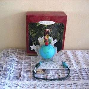 May include: A clear plastic Christmas ornament with a light blue globe and a small figure of a person playing a trumpet. The ornament is lit up and has a wire cord. The ornament is in its original box, which is red with a white label that says "Keep Sake Ornament".