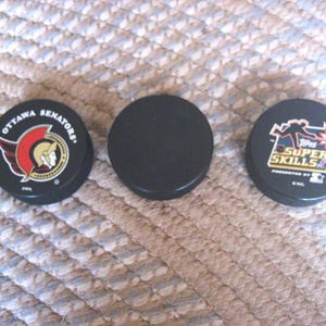 May include: Three black hockey pucks. Two pucks have graphics: one with the Ottawa Senators logo and the other with "Super Skills" text. The third puck is plain black. All are on a textured surface.