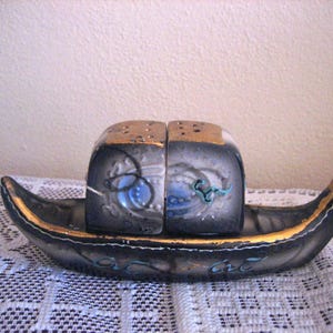 May include: A vintage salt and pepper shaker set in the shape of a gondola. The set features a black base with gold trim and two salt and pepper shakers with a blue and gold design. The shakers have a gold top with holes.