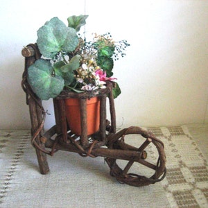 May include: A small, rustic wooden wheelbarrow planter with a brown terracotta pot filled with green leafy plants and pink flowers.
