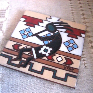 May include: Square ceramic tile featuring a Kokopelli figure playing a flute, in black, white, and turquoise. The tile has a beige background with geometric patterns in red, brown, and turquoise. The tile is approximately 4 inches square.