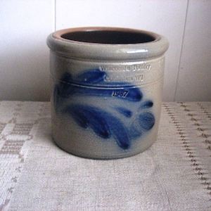 May include: A stoneware crock with a blue floral design. The crock has a light gray base with a dark brown rim. The text "Wisconsin Pottery Columbus WI 1987" is printed on the side.