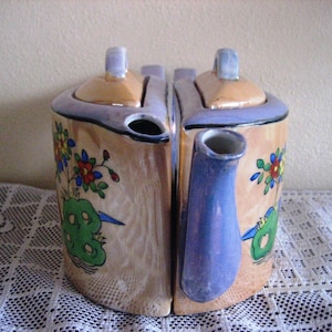 May include: A vintage ceramic teapot with a unique double-sided design. The teapot features a tan base with floral and bird motifs, a blue spout, and light blue accents. The lids are peach-colored with light blue handles.