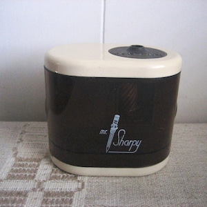 May include: A vintage black and cream-colored pencil sharpener with the text "mr. Sharpy" in a cursive font. The sharpener has a rounded rectangular shape with a small, circular opening on top.
