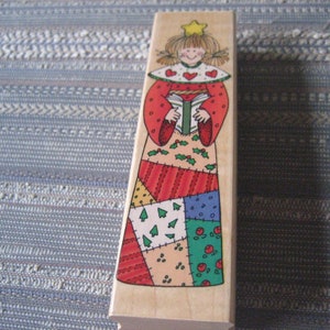 May include: A wooden Christmas stamp featuring a patchwork angel holding a book. The angel has a red dress, a white collar with red hearts, and a gold star on her head.