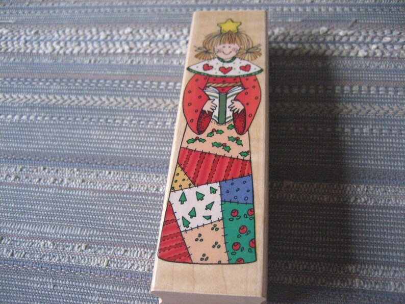 May include: A wooden Christmas stamp featuring a patchwork angel holding a book. The angel has a red dress, a white collar with red hearts, and a gold star on her head.