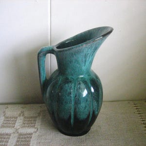 May include: A ceramic pitcher featuring a deep teal and dark green mottled glaze with a glossy finish. The vessel has a rounded, ribbed body, a curved handle on the side, and a wide, elongated pouring spout extending from the top rim.