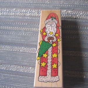 May include: A wooden Christmas stamp featuring a red and white Santa Claus holding a green Christmas tree. The stamp has a white border and is decorated with yellow stars.