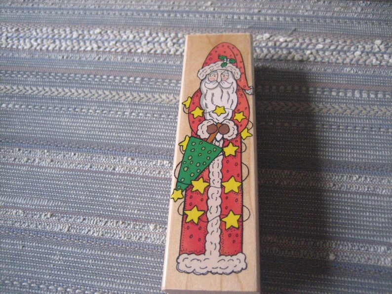 May include: A wooden Christmas stamp featuring a red and white Santa Claus holding a green Christmas tree. The stamp has a white border and is decorated with yellow stars.