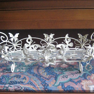 May include: A white metal decorative tray with a butterfly and floral design. The tray has a rectangular shape with curved handles and is supported by four small, curved feet. The tray is placed on a patterned fabric.