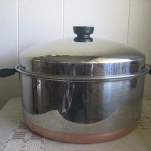 May include: A large, round, stainless steel stockpot with a polished finish, featuring a copper-colored base, two black side handles, and a matching stainless steel lid with a black knob handle centered on top for secure coverage.
