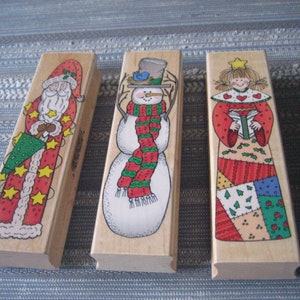 May include: Three wooden Christmas stamps featuring a snowman, Santa Claus, and an angel. The snowman is wearing a red and green scarf and a blue hat. Santa Claus is holding a green and red Christmas tree. The angel is wearing a red and green dress with a patchwork skirt and holding a book.