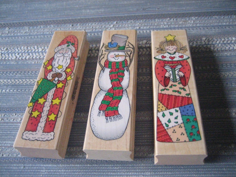 May include: Three wooden Christmas stamps featuring a snowman, Santa Claus, and an angel. The snowman is wearing a red and green scarf and a blue hat. Santa Claus is holding a green and red Christmas tree. The angel is wearing a red and green dress with a patchwork skirt and holding a book.
