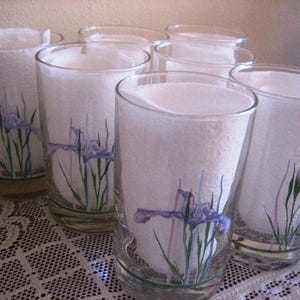 May include: Set of six clear drinking glasses. Each glass features a hand-painted design of purple iris flowers and green stems. The glasses are filled with white paper and are arranged on a white lace tablecloth.