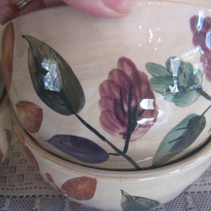 May include: Cream-colored ceramic bowls with a floral design. The bowls are stacked, showcasing a hand-painted pattern of green, purple, and brown leaves and accents. The bowls have a glossy finish and a slightly irregular shape.