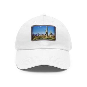 Peut inclure: Une casquette de baseball blanche avec un écusson rectangulaire sur le devant. L'écusson représente un paysage désertique avec de grands cactus saguaro, de la végétation verte, des montagnes au loin et un ciel bleu avec un nuage fin.