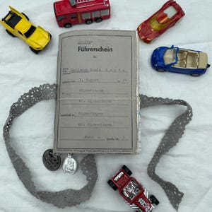 Könnte beinhalten: Eine Sammlung von Spielzeugautos in Rot, Gelb und Blau, zusammen mit einem alten deutschen Dokument und zwei kleinen Silberanhängern. Das Dokument ist mit einem grauen Spitzenband gebunden. Die Autos umfassen einen Feuerwehrauto, ein Cabrio und einen Hot Rod.