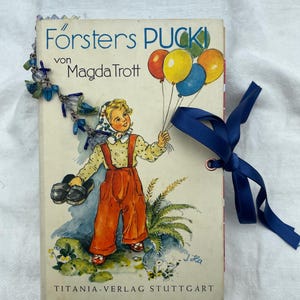 Könnte beinhalten: Ein gebundenes Vintage-Buch mit dem Titel Försters PUCK von Magda Trott, das ein Kind mit Luftballons zeigt. Das Buch ist mit einem blauen Band, das durch eine Metallöse gezogen ist, und einer Perlenkette mit blauen Anhängern verziert.