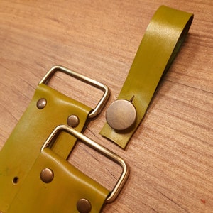 May include: Two olive green leather straps with brass buckles and a brass button. The straps are connected to each other with a brass buckle.