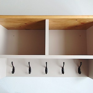 May include: A white wooden wall shelf with two open compartments and a row of five black metal hooks underneath. The top of the shelf is made of natural wood.