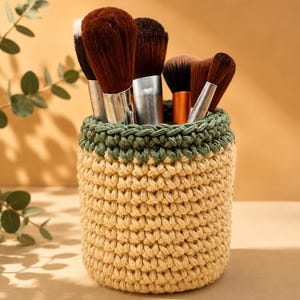 May include: A handmade crochet makeup brush holder in beige and olive green. The holder contains several makeup brushes with brown bristles and silver handles. The crochet design is visible, showcasing the texture and craftsmanship of the piece.