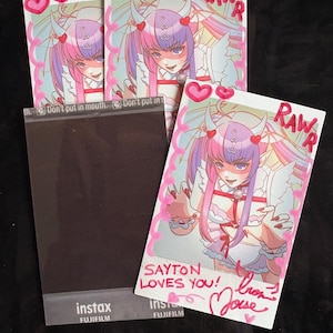 May include: Several Instax photos featuring a cartoon character with pink and purple hair, horns, and angel wings. The photos have the text "RAWR" and "SAYTON LOVES YOU!" written on them. The photos are on a dark surface.