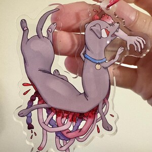 May include: A clear acrylic keychain featuring a cartoon dog with a blue collar and a gold tag. The dog is depicted with a hand in its mouth and tentacles with red and purple accents. The keychain has a red metal ring.