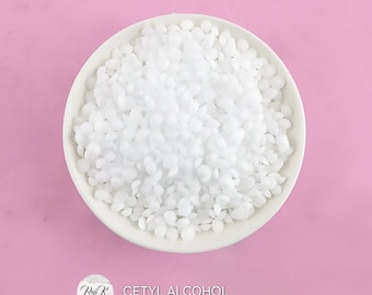 May include: A white bowl filled with white cetyl alcohol flakes. The bowl is on a pink surface. The text "Cetyl Alcohol" is written below the bowl.