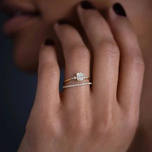 Puede incluir: Un anillo de oro con un diamante de forma cuadrada y diamantes más pequeños en la banda. El anillo se lleva en el dedo anular de la mano izquierda de una persona. Una banda delgada de oro con pequeños diamantes se lleva en el dedo debajo del anillo principal.