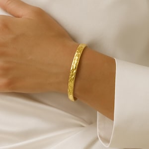 May include: A gold-coloured bangle bracelet with a hammered texture. The bracelet is approximately 6.35 cm in diameter and is worn on a wrist. The background is a white shirt.