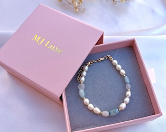 May include: A delicate bracelet with white pearls and light blue stones, displayed in a pink box with the text "MJ Luxe" on the lid.