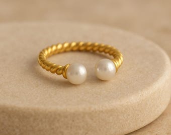 Può includere: Un anello dorato con una fascia intrecciata e due accenti di perle bianche.
