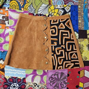 May include: A brown suede mini skirt with a unique design. The skirt features a scalloped edge with button closures and a contrasting panel with a black and brown geometric pattern. The skirt is displayed on a colorful patchwork background.