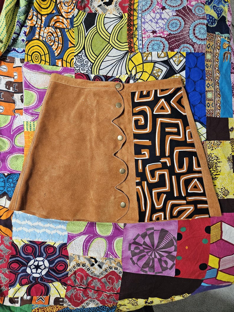 May include: A brown suede mini skirt with a unique design. The skirt features a scalloped edge with button closures and a contrasting panel with a black and brown geometric pattern. The skirt is displayed on a colorful patchwork background.