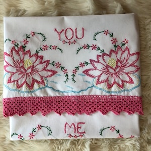 May include: White embroidered fabric with pink lotus flowers, green vines, and the words "YOU" and "ME". A pink crocheted lace border adds detail. The fabric is folded.