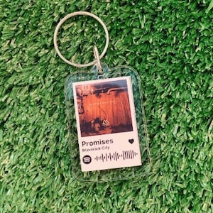May include: A clear keychain featuring a photograph of a building with a red and orange facade. The text "Promises" and "Maverick City" are printed on the keychain. A Spotify barcode is also printed on the keychain.