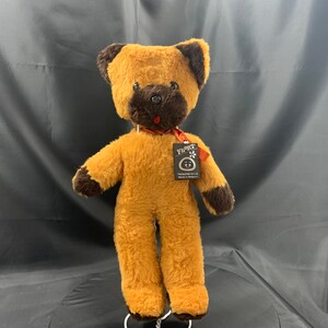Puede incluir: Un oso de peluche marrón y beige con la cara negra y una cinta roja alrededor del cuello. El oso está de pie sobre una superficie blanca y tiene una etiqueta que dice "¡Yippee!"