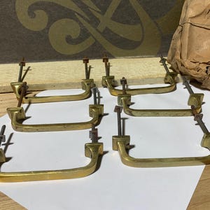 Könnte beinhalten: Sechs Vintage-Messing-Schubladengriffe mit Befestigungsmaterial sind auf einer weißen Oberfläche angeordnet. Die Griffe haben ein gebogenes Griffdesign und sind goldfarben. Ein in braunes Papier gewickeltes Bündel befindet sich im Hintergrund.