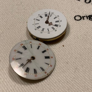 Puede incluir: Dos esferas de reloj antiguas con marcadores de hora en números romanos y agujas negras. Las esferas son blancas con ribetes dorados y se encuentran sobre un fondo de tela blanca.