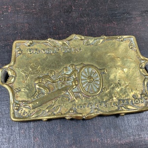 May include: Antique brass plaque with a detailed relief depicting two figures pushing a cannon. The plaque has decorative floral edges and the text "UN VRAI FOLU NOTRE 75 ACTION".