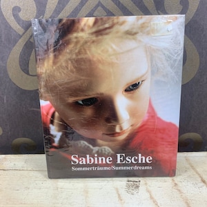 Puede incluir: Un libro titulado "Sabine Esche Sommerräume/Summerdreams" con una imagen en primer plano de una persona con piel clara y cabello claro. La persona lleva una parte superior roja. El libro está envuelto en plástico transparente.